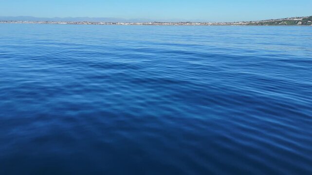 Aerial drone shot flying over calm blue ocean surface with shimmering sunlight reflection and gentle ripples, cinematic seascape background in natural daylight