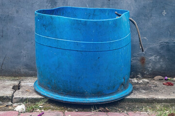 Obraz premium A weathered blue plastic barrel with a bent rusty metal handle is positioned outdoors on a cracked concrete surface, against a rough textured concrete wall