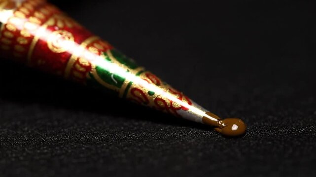 A decorative henna cone lies on a dark surface, intricately designed with red and green patterns, awaiting application.