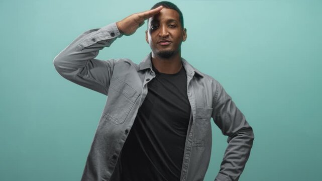 Young black man salutes with right hand to forehead, hand on hip, wearing gray shirt and black tee in studio with teal backdrop; confidence respect.