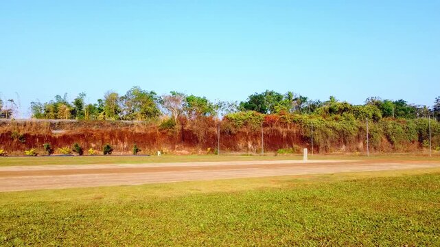 A wide, panning shot of a professional cricket pitch and field under a clear blue sky, featuring white crease markings, a sightscreen, and a lush green outfield bordered by tropical trees.