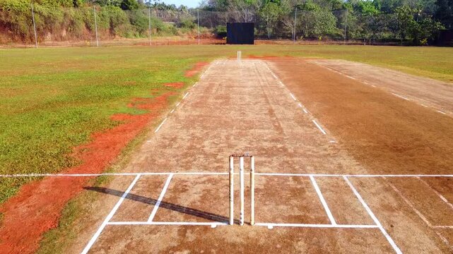 Aerial drone shot of a rural cricket pitch and wickets on a sunny day, showcasing the dirt crease, green outfield, and surrounding tropical trees in a peaceful sports field setting.