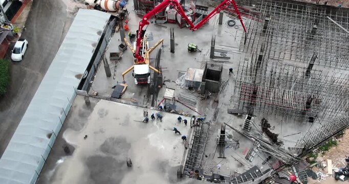 Aerial view of a construction site during rain, featuring a truck with a boom pumping concrete onto a building foundation as workers level the wet cement beside  an intricate grid of scaffolding.