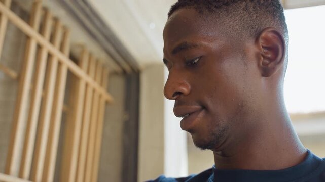 Black man repairing house window frame, sweaty handyman inspecting wooden slats, measuring and tightening screws inside sunlit residential interior, ladder and toolbox visible, focused expression,