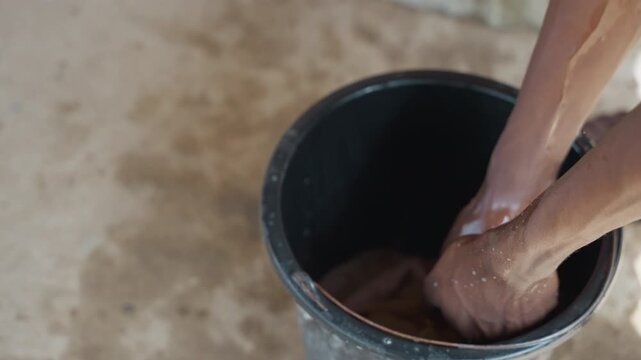 Hands washing clothes in black bucket, closeup of soapy palms scrubbing fabric over concrete floor, sudsy water splashing and being wrung out, outdoor household laundry chore conveying quiet effort