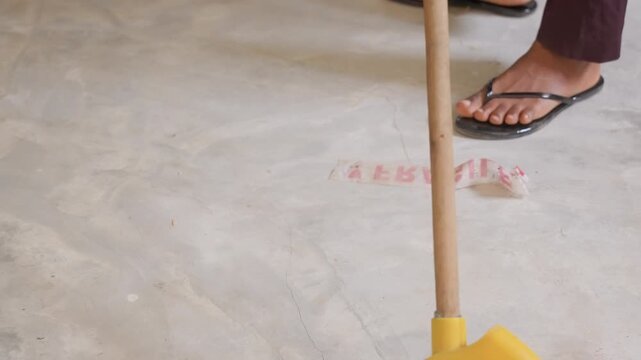 Pink mop sweeping house concrete floor with yellow handle, sandals stepping nearby, dust and residue gathered, casual cleaning motion and rhythmic sweep suggested plot homeowner performing daily