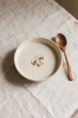 Obraz premium Creamy White Mushroom Soup Elegance: Top view of creamy white mushroom soup in a matte ivory bowl, styled on a linen tablecloth with soft beige tones, wooden spoon on the side.