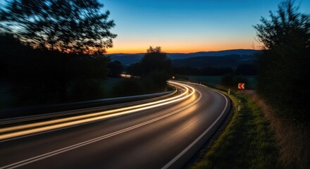A winding road at dusk with blurred headlights and trees on either side.