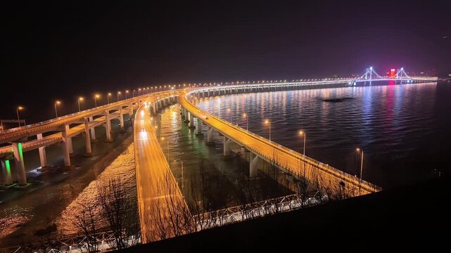 Timelapse of Xinghai Bay Bridge illuminated at night in Dalian, China. Long exposure captures dynamic traffic light trails across the sea-spanning bridge, with city skyline glowing in the background 