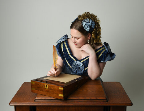 portrait of Victorian Era Woman in historical victorian classical Blue Ball Gown costume, Plus Size Model. sitting at antique letter writing desk holding quill. isolated figure on studio background.
