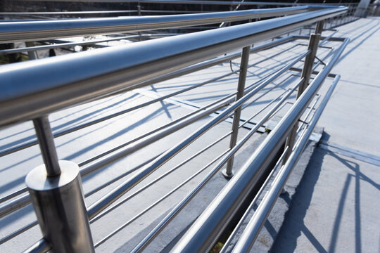 Perspective view of a polished stainless steel railing and handrail on a modern walkway, showing tubular bars, joints, reflections and shadow patterns that emphasize minimalist industrial architecture