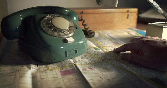 Close up of a hand identifying a location on a street map before dialing a vintage phone. Concept for tracking suspects or police coordination.