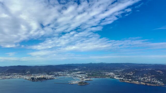 An elevated aerial 4K view of Ibiza (IBZ) city and harbor taken from an airplane cockpit in a sunny day with few clouds in a blue sky.