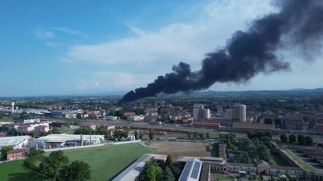 Dense black smoke plume rising above urban district with railway yard and residential building in background, spreading across blue sky over cityscape and surrounding landscape, drone static aerial sh