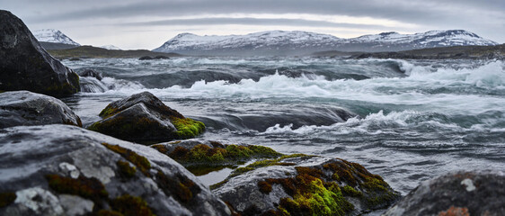 Ultrawide Landscape of a Rushing Mountain River with White Water Rapids, Mossy Rocks in the Foreground, and Snow-Capped Peaks under a Moody Overcast Sky in a Wild Northern Wilderness Setting