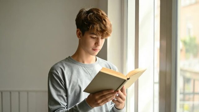 Young man engrossed in reading a book by a bright window, enjoying quiet study time