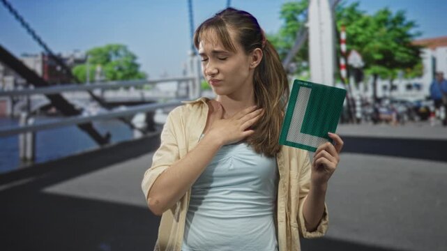 Young woman holding learner plate and pressing hand to throat on a busy street bridge near canal, visible discomfort and learner sign; learner stress.