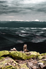 Vertical cliffs give way to gentle slopes and a cairn balances on lichen stone as mist floats over the valley. Snow peaks line the horizon and the mood is calm and adventurous