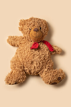 a well loved fluffy brown teddy bear with a red scarf lying down on a plain background