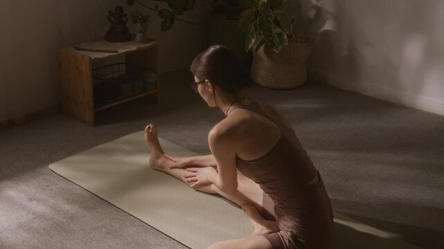 Full rear shot of young woman in beige sports top and sorts doing janu shirshasana, bending towards extended leg, focusing on correct posture and technique, during home yoga practice
