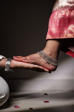 Close-up of an Indian bride&rsquo;s decorated foot with henna and silver anklet during a traditional wedding ritual, groom holding her foot under red lighting.