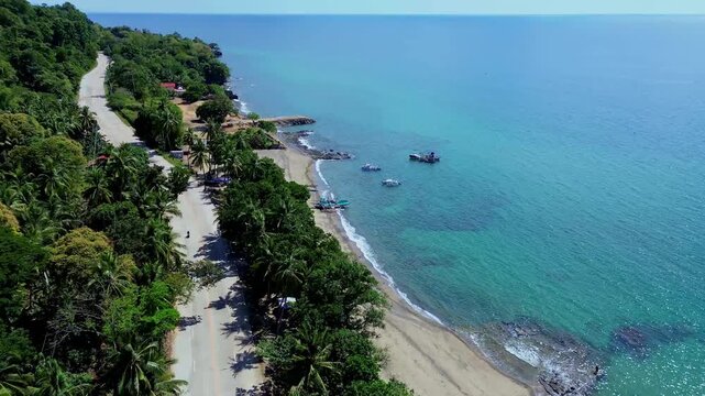 Aerial view drone fly over to the scenic a coastal road bordered by lush greenery and a calm blue sea with moored outrigger boats showing travel experience in Basay, Negros Oriental, Philippines