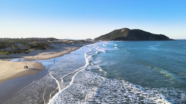 Cinematic aerial pull-back shot of Santinho Beach in Florianopolis, Brazil, revealing turquoise ocean waves, white sand dunes and the Morro dos Ingleses mountain under a clear blue sky.