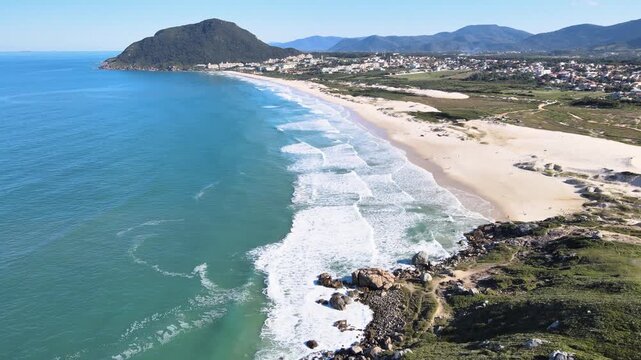 Cinematic aerial panorama of Santinho Beach in Florianopolis, Brazil, showcasing turquoise ocean waves, white sand dunes and the lush Morro das Aranhas mountain under a clear blue sky.