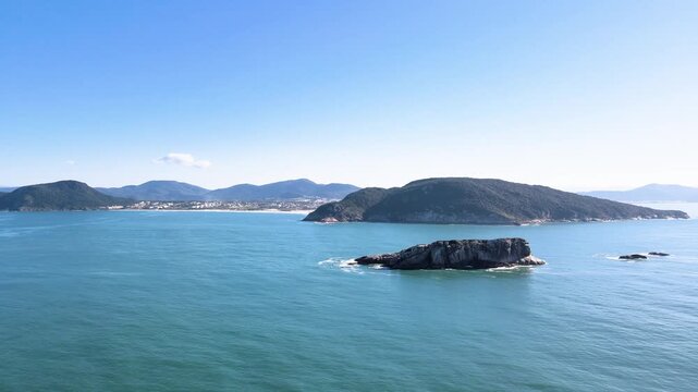 Cinematic aerial panorama of Ilha do Badejo in Florianopolis, Brazil, featuring rocky formations in the Atlantic Ocean with Santinho Beach and Morro dos Ingleses in the background.