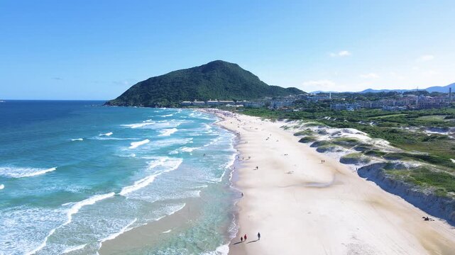 Cinematic aerial wide shot of Santinho Beach in Florianopolis, Brazil, featuring turquoise ocean waves, white sand shoreline and the lush Morro das Aranhas mountain under a clear blue sky.