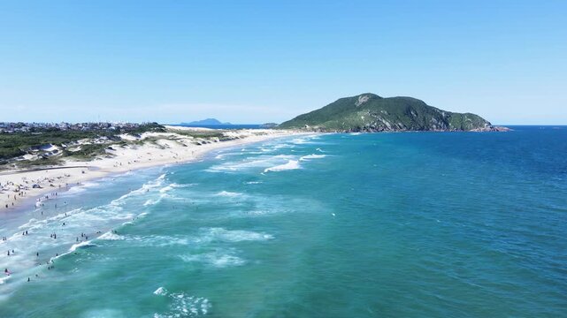 Cinematic aerial wide shot of Santinho Beach in Florianopolis, Brazil, showcasing turquoise ocean waves, white sand dunes and the Morro dos Ingleses mountain under a clear blue sky.
