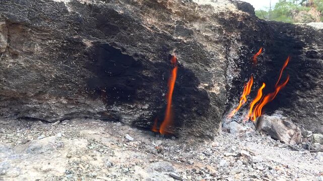 Natural methane gas fire burns between rocks on barren ground across a rugged rocky landscape. Eternal flames rise from stone gaps as subsurface fuel ignites within raw desolate terrain.