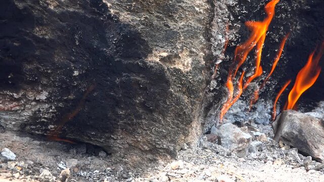Natural methane gas fire burns between rocks on barren ground across a rugged rocky landscape. Eternal flames rise from stone gaps as subsurface fuel ignites within raw desolate terrain.