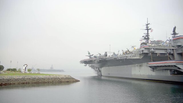 Wide shot of the USS Midway aircraft carrier museum docked in San Diego Bay with the Unconditional Surrender statue in the foreground. Iconic waterfront landmark and naval history attraction in Califo