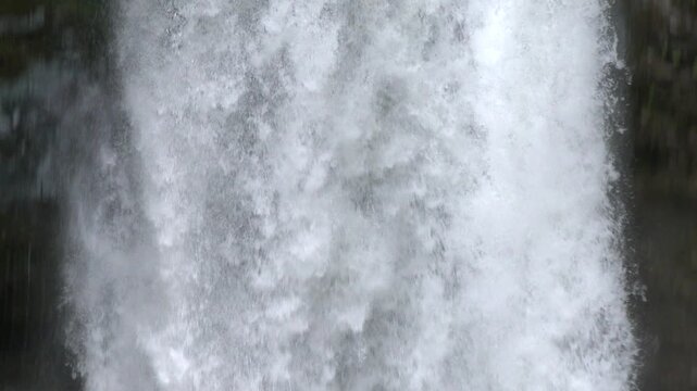 Camera follows powerful waterfall water flowing through dense green forest captured in super slow motion. Rushing cascade and mist reveal dramatic natural energy as falling water fills the frame.