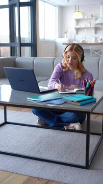 Lovely concentrated blonde teenage girl in wireless headphones e-learning using laptop pc, studying online lesson and writing assignment into exercise book in domestic room.