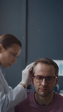 Vertical eye level shot of female dermatologist wearing gloves examining male patient scalp during dermatology consultation in clinic, professional skin examination procedure in medical office