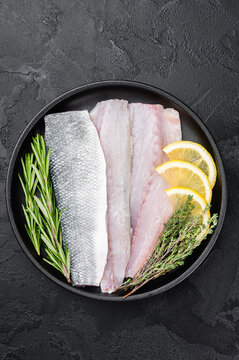 Uncooked European sea bass fillet on black plate with rosemary and thyme, clean culinary layout ideal for seafood menu, cooking recipe or nutrition design.