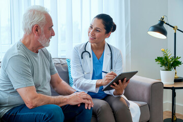 Obraz premium Caucasian senior man sitting on sofa talking with Hispanic female doctor holding digital tablet, medical professional listening attentively and taking notes during consultation