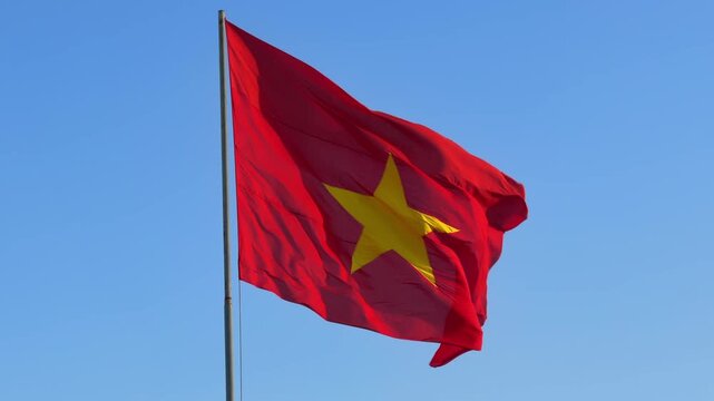 Slow motion red Vietnam flag with yellow star waves strongly in the wind, glowing in warm sunset light against a clear blue evening sky above the landscape. National pride.