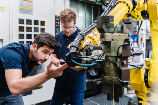 Male coworkers examining robotic arm while standing in manufacturing factory