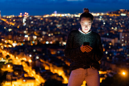 Young woman using cell phone at night above the city, Barcelona, Spain