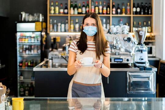 Beautiful owner wearing mask while holding coffee cup and saucer in cafe