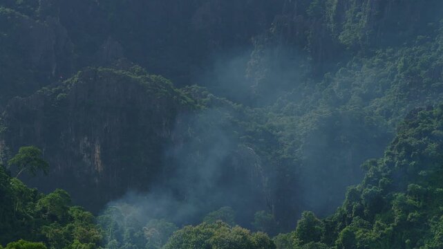 Smoke of slash and burn fire is risinng from the trees among the mountains, with upward lifting movement view with moutainous tripical landscape in the background.