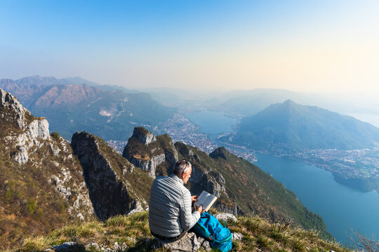 Rear view of hiker reading a book on mountaintop, Orobie Alps, Lecco, Italy