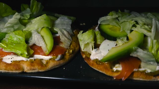 Close sliding view of nutritious open sandwiches topped with smoked salmon avocado egg and greens on rustic oat Lapland bread. Focus on highlighted appetizing fresh textures arranged on black plate