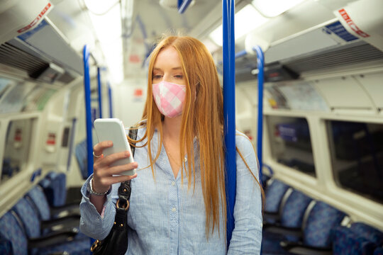 Young woman in mask using smart phone while standing in train