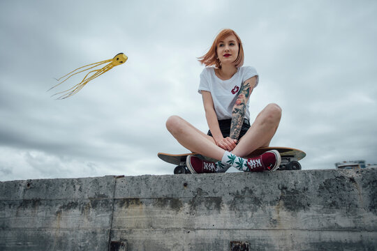 Young woman sitting on a concrete wall on carver skateboard with kite in background