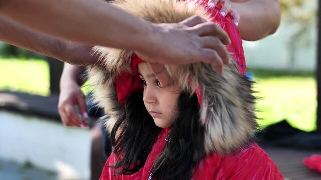 Mother helping young Armenian girl putting on a red winter coat with fur-lined hood.