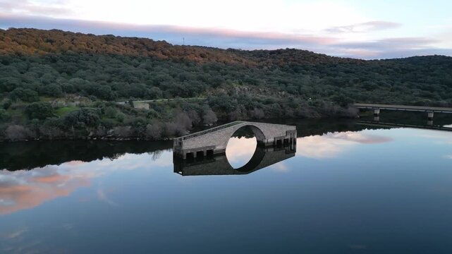 Aerial drone view of the Roman Bridge of El Ponton at sunset in Guijo de Granadilla, Extremadura. Warm golden light over historic stone structure.